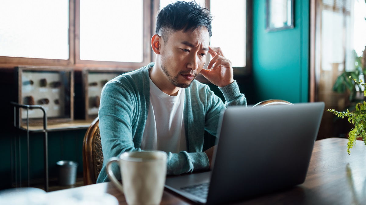 asian man at laptop on a desk looking stressed with hand on forehead